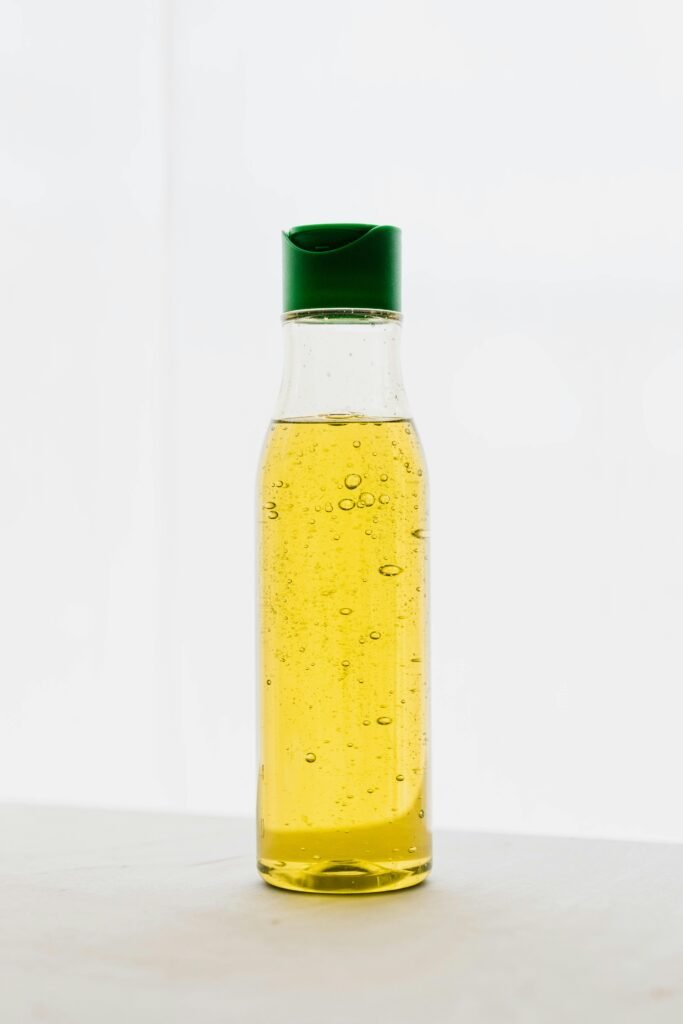 Yellow body oil with bubbles in transparent plastic bottle with green cap on table against white background